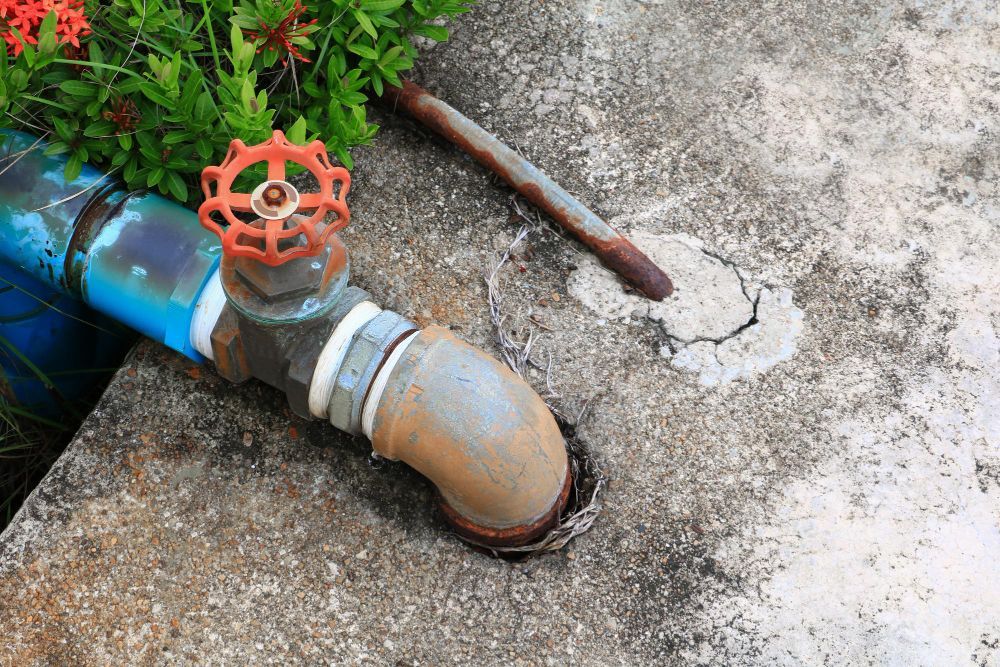 A bright orange gate valve attached to a blue pipe and a rusted metal elbow, set against a concrete background.