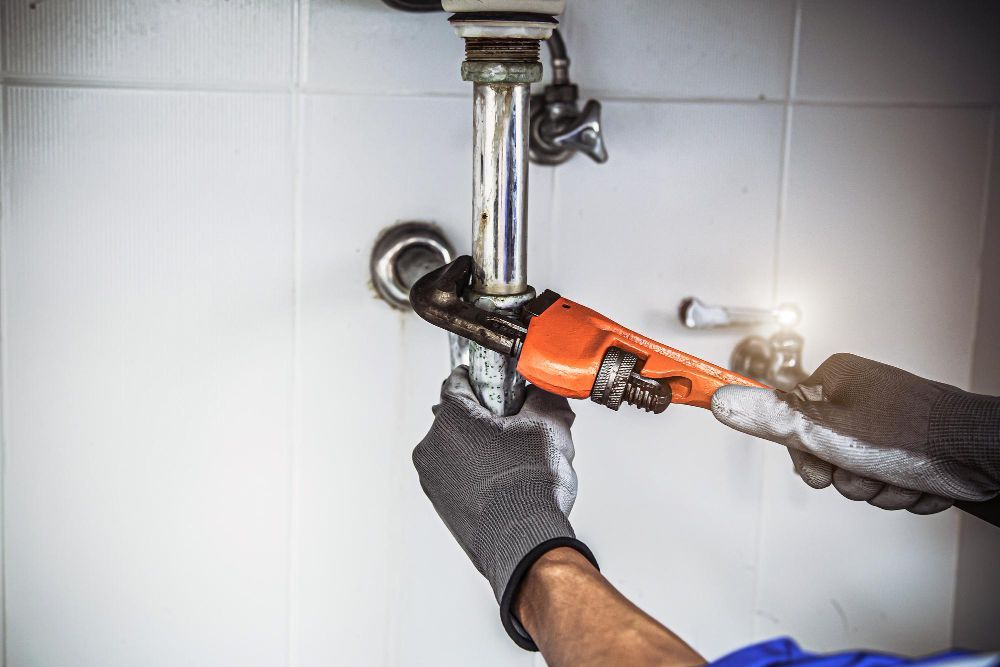 A person in grey work gloves uses an orange pipe wrench to tighten a metal plumbing pipe against a white tiled wall.