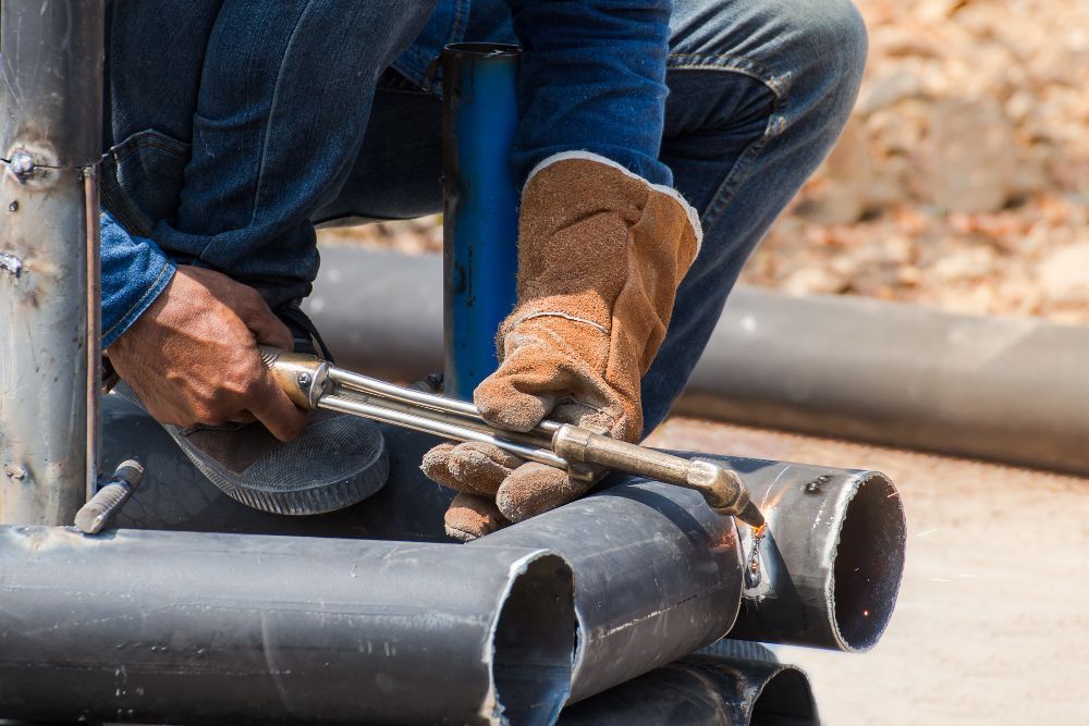 A person wearing tan leather gloves uses a torch to cut through a metal pipe outdoors.