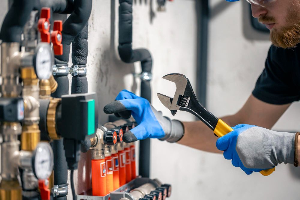 A technician wearing blue gloves uses an adjustable wrench to adjust valves on a home heating system.