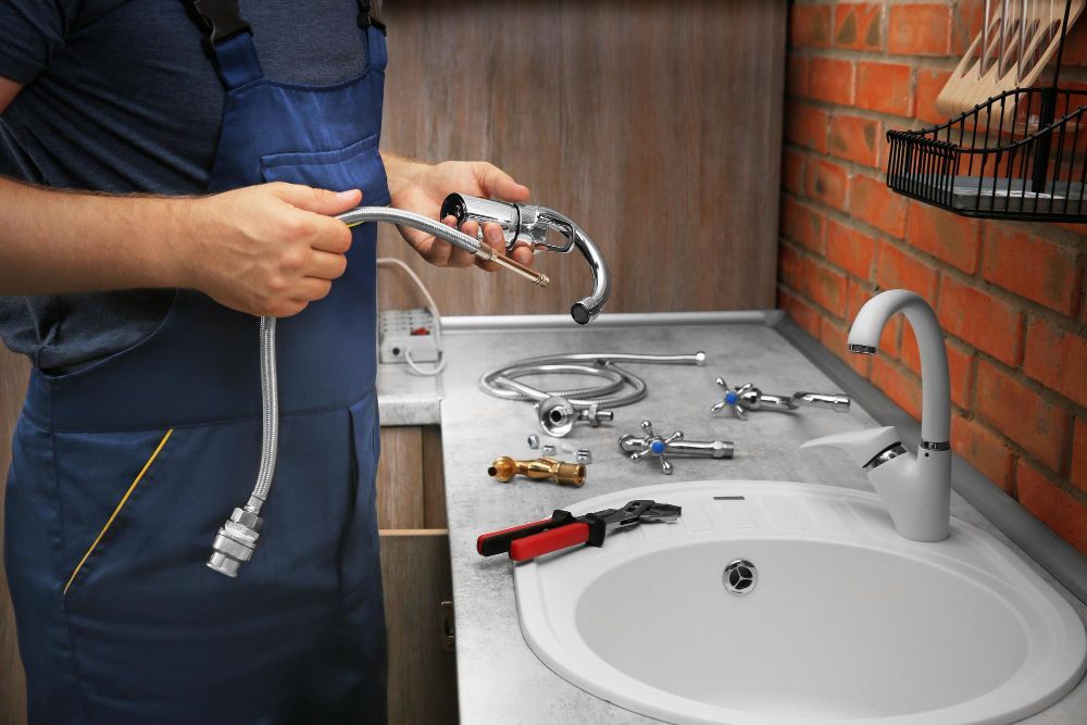 A plumber in blue overalls holding faucet parts above a kitchen sink, with tools and components laid out on the counter.