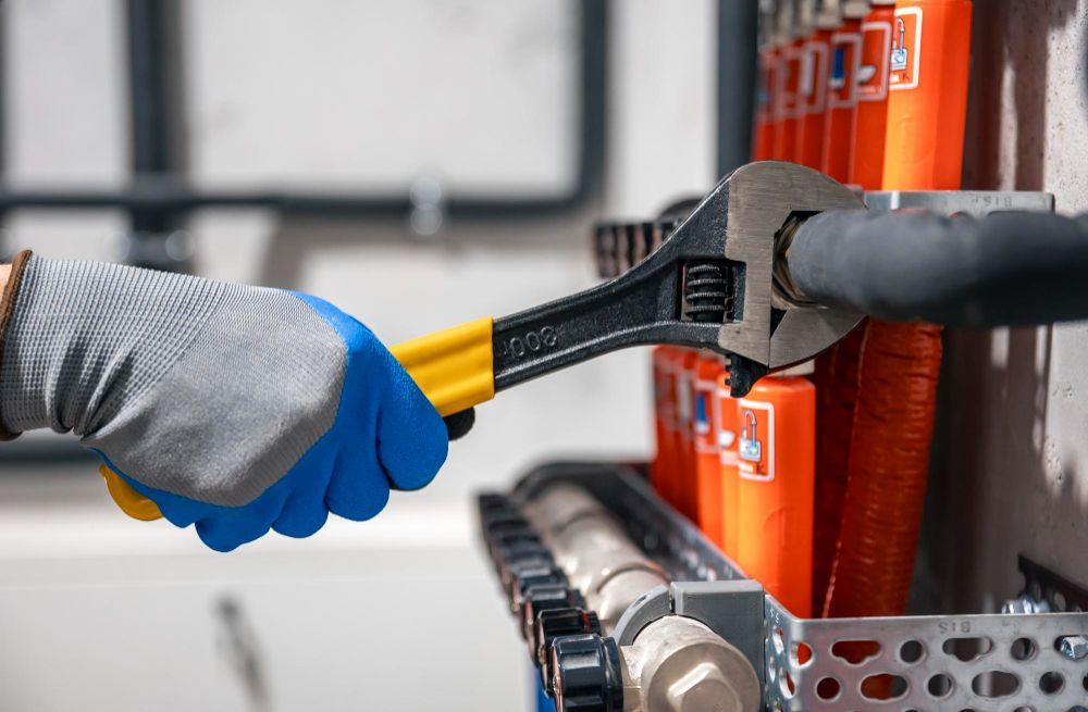 A gloved hand uses an adjustable wrench to tighten a fitting on a mechanical manifold system with orange labels.