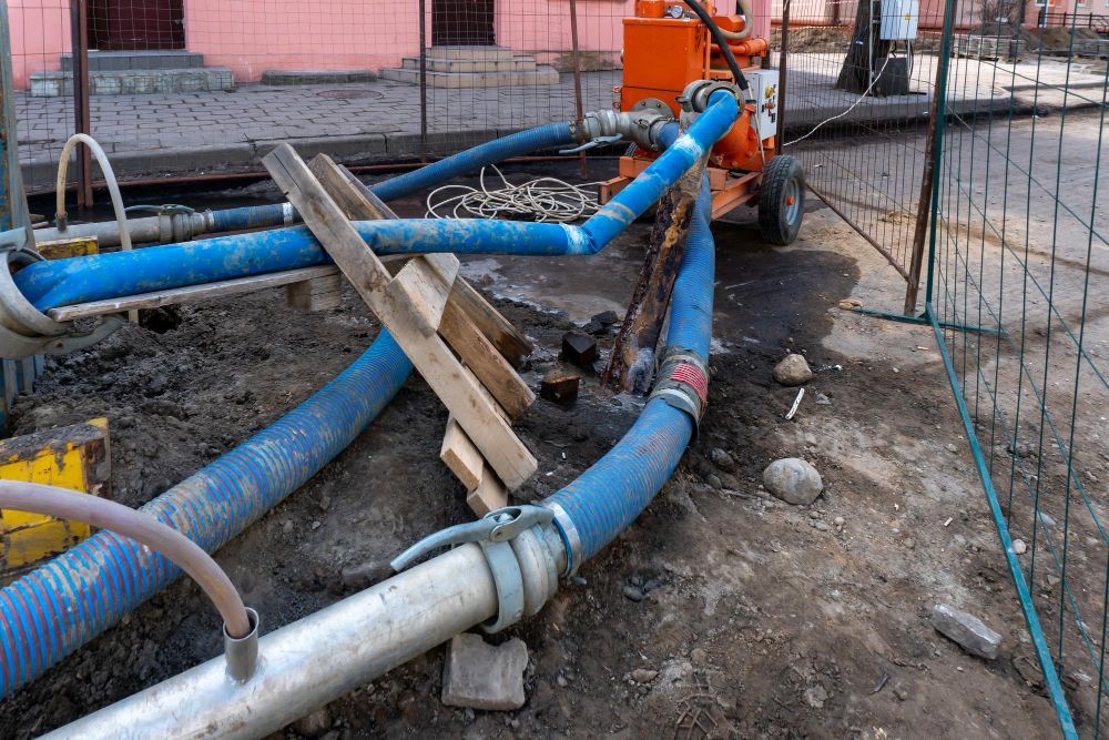 Construction site with orange machinery and blue water-pumping hoses connected to pipes on muddy ground.