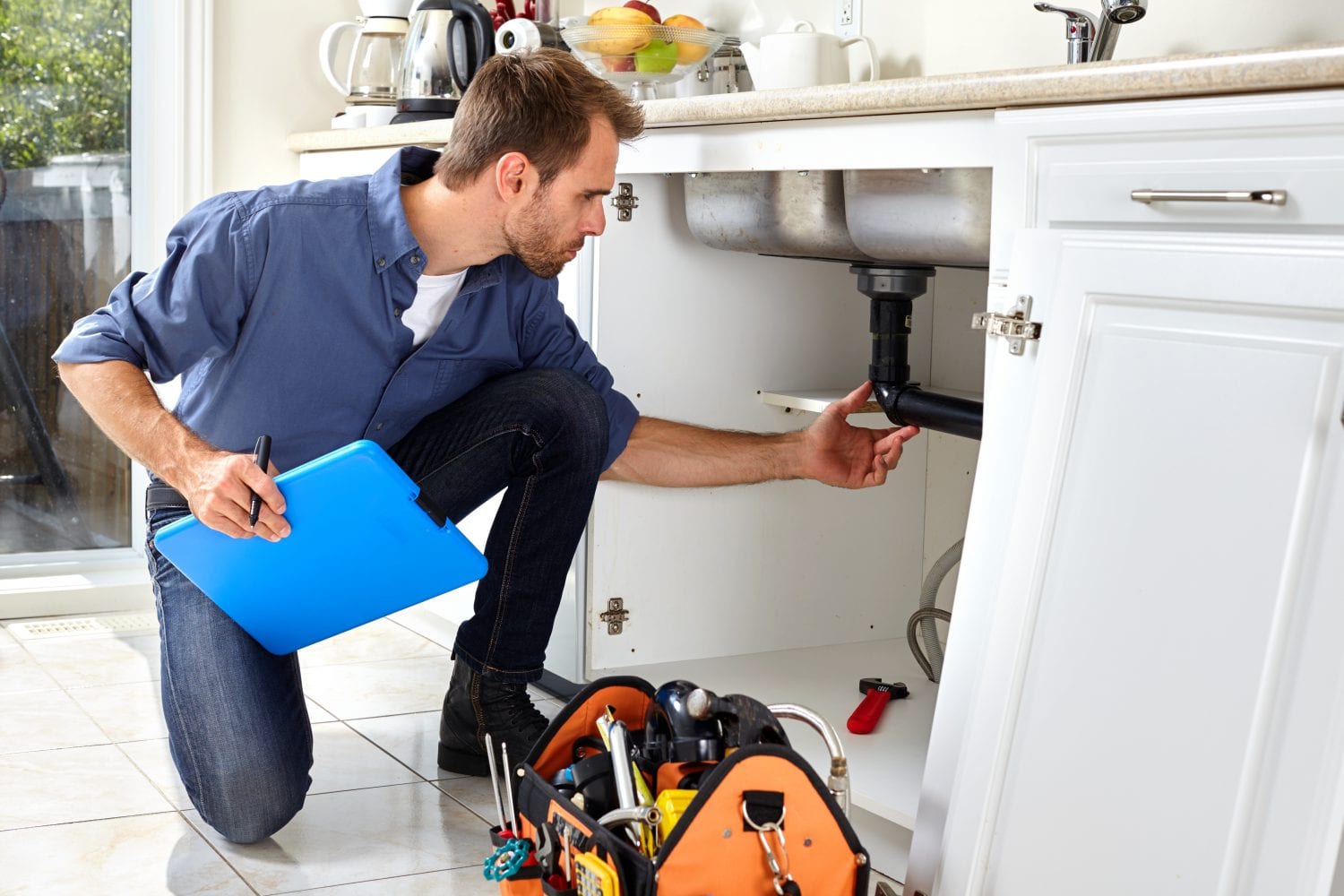 A plumber is kneeling down in a kitchen looking under a sink.