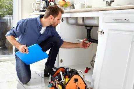 A technician holding a blue clipboard inspects plumbing pipes under a kitchen sink, with a tool bag nearby.