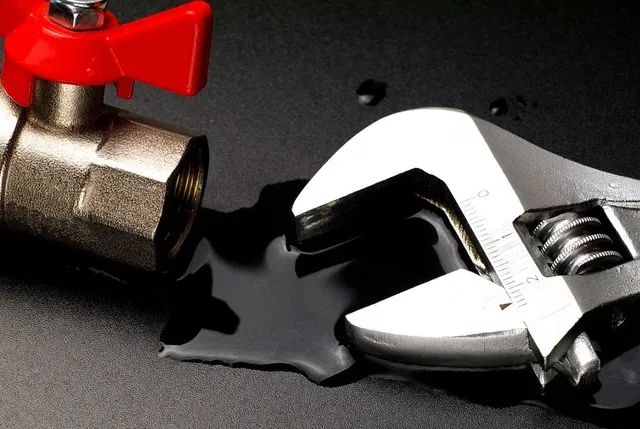 An adjustable wrench lies next to a metal ball valve on a dark surface with a puddle of liquid.
