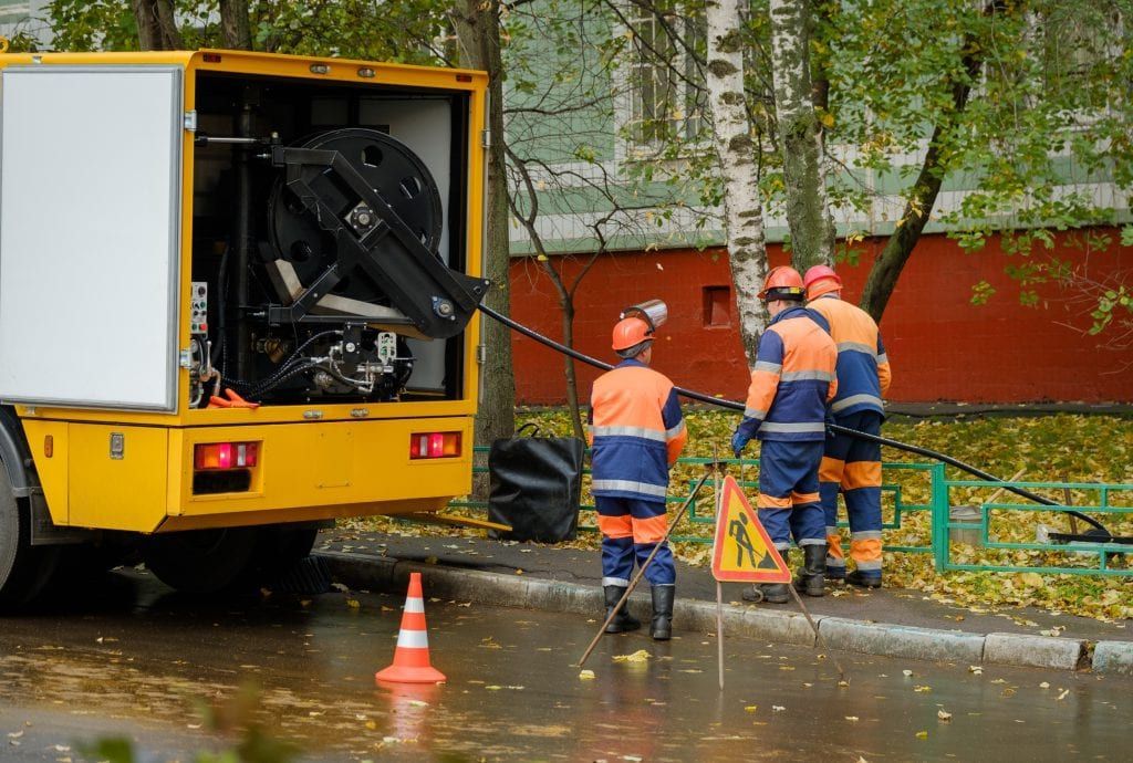 Three workers in orange and blue uniforms work near a yellow utility truck on a wet, leaf-covered street.