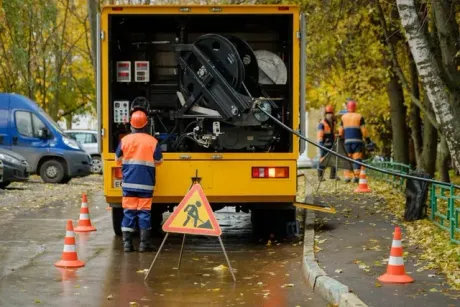 Utility workers in high-visibility gear work near a yellow maintenance truck with traffic cones on a tree-lined street.