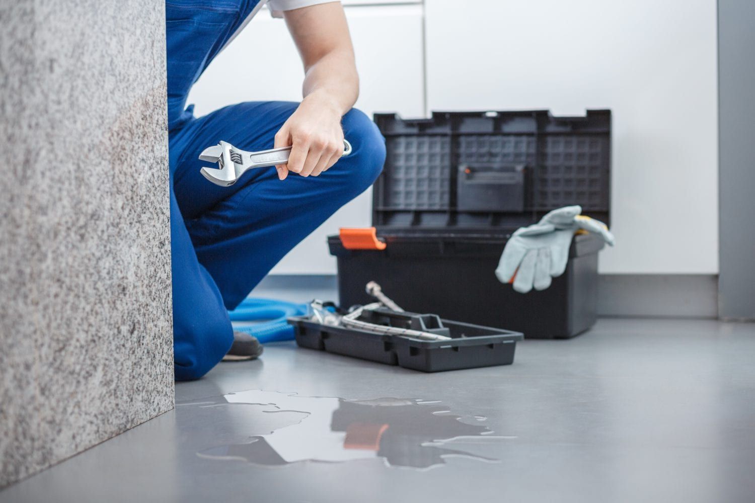 A plumber in blue overalls kneeling by a water leak on the floor with a wrench and toolbox nearby.