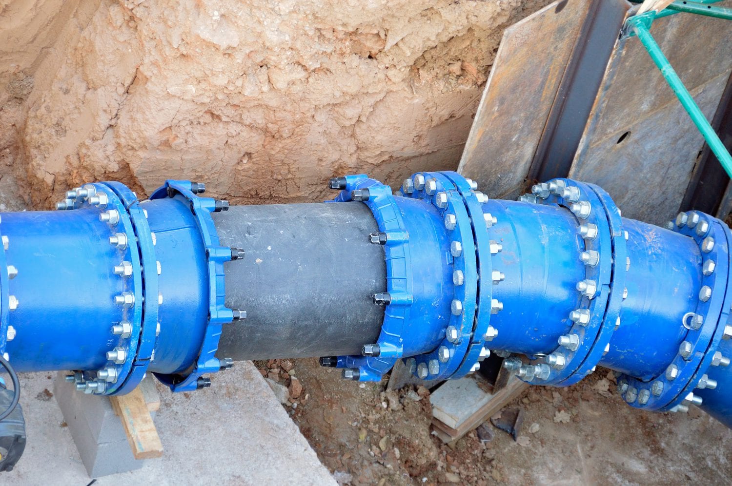 A blue industrial pipe section connected with flanged bolts and a black center sleeve in an excavated dirt trench.