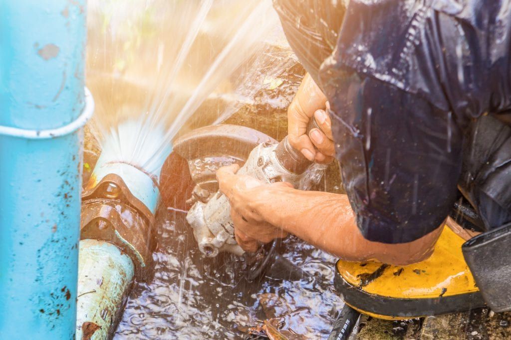 A worker in dark clothing and yellow boots uses a power tool on a leaking blue pipe spraying water.