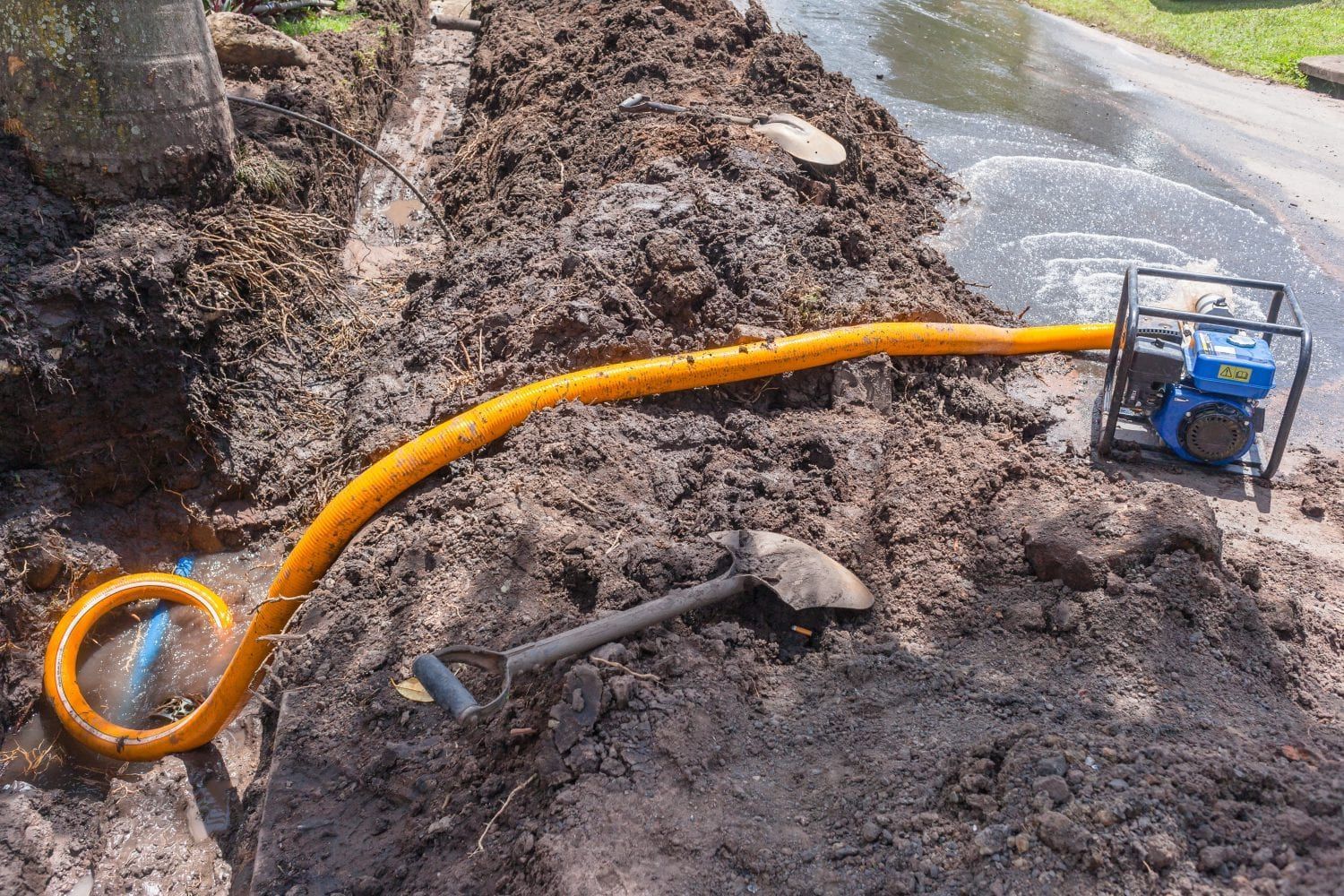 A portable yellow water pump in a muddy trench is draining water onto a nearby road, with a shovel lying in the dirt.