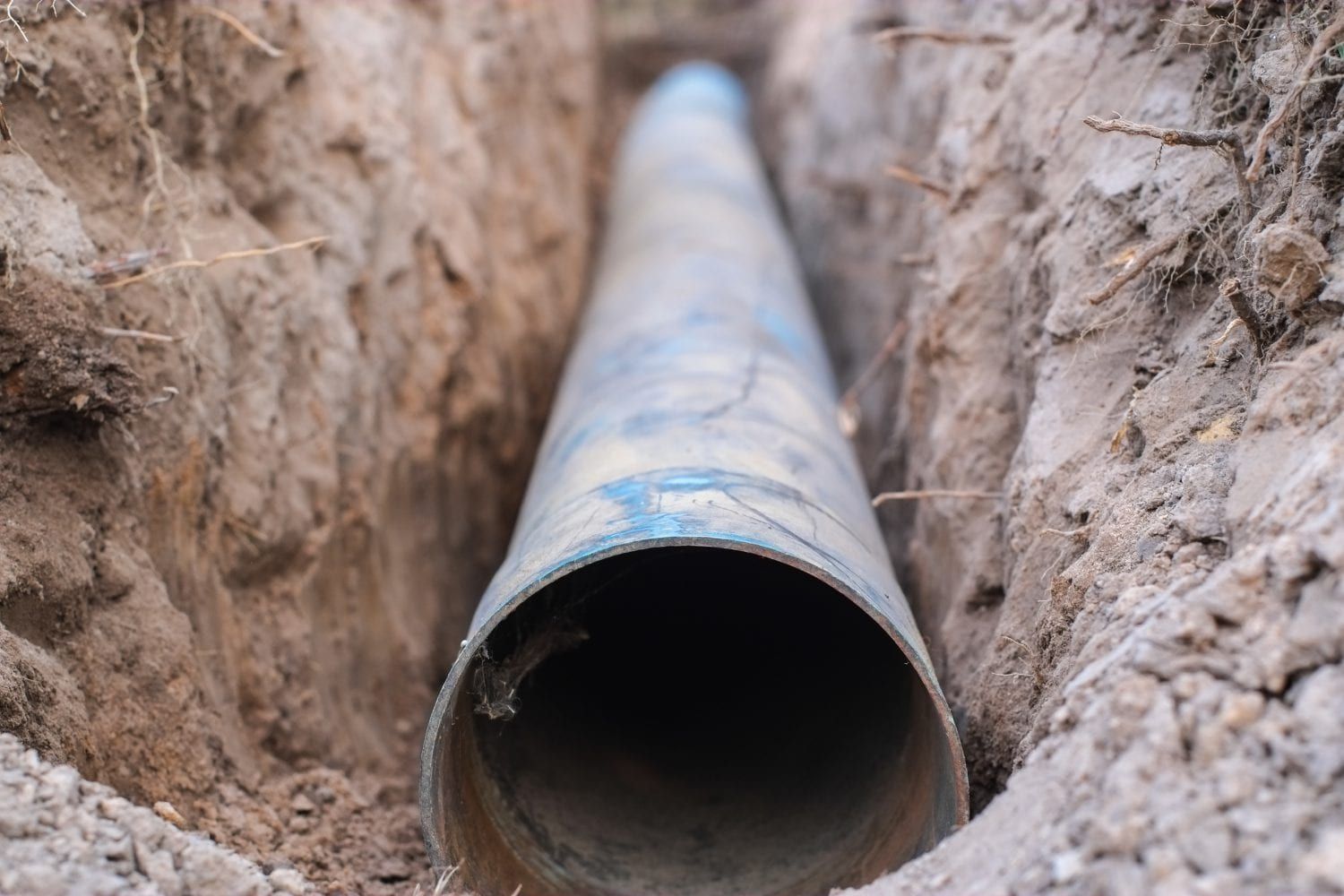 A long, cylindrical blue pipe lies at the bottom of a narrow, freshly dug dirt trench.