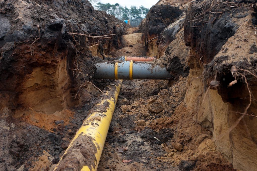 A deep trench shows a long yellow pipe running lengthwise with a gray T-shaped pipe spanning across it.