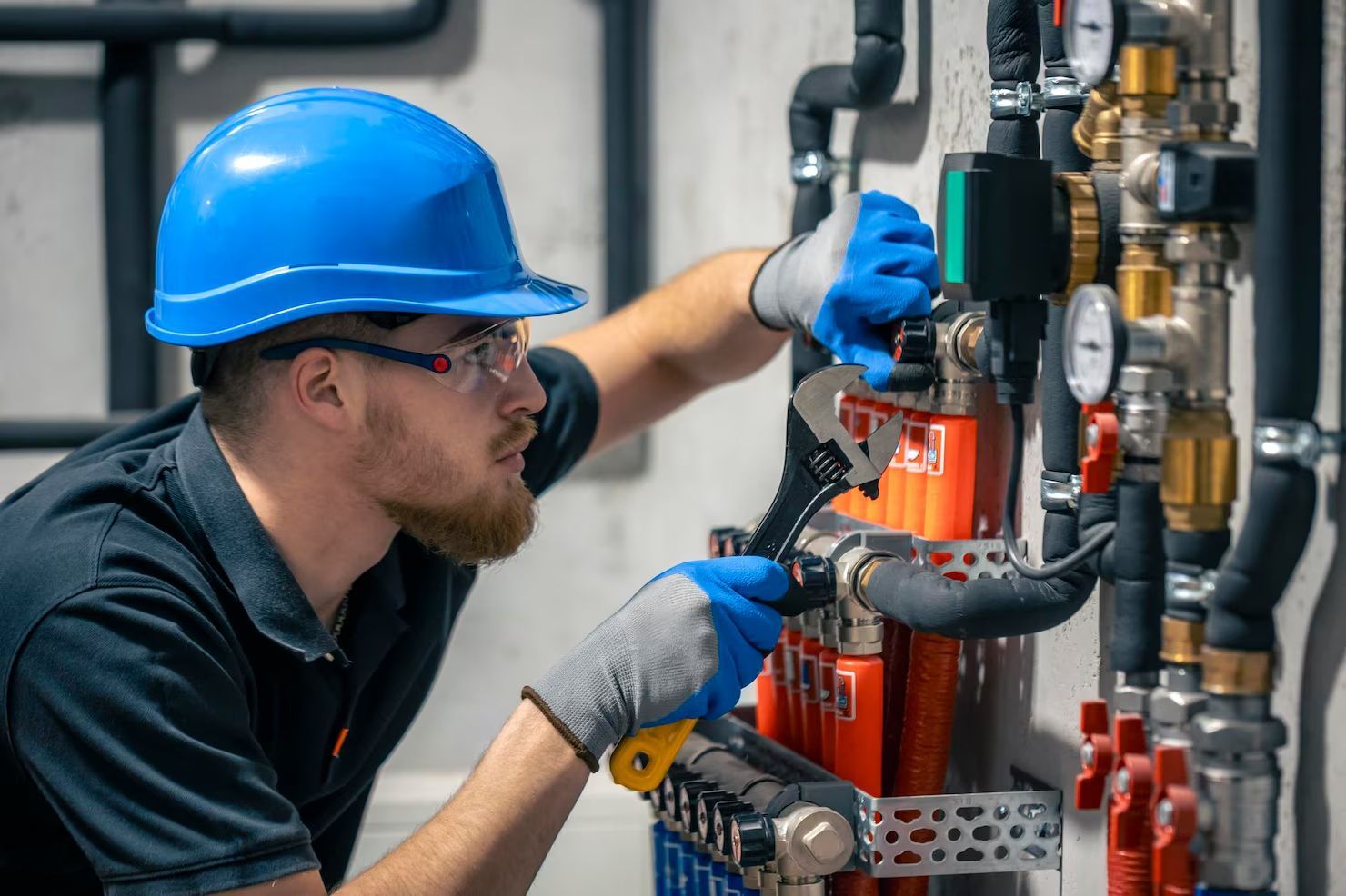 A worker wearing a blue hard hat and gloves uses a wrench to adjust orange pipes in a mechanical or utility room.