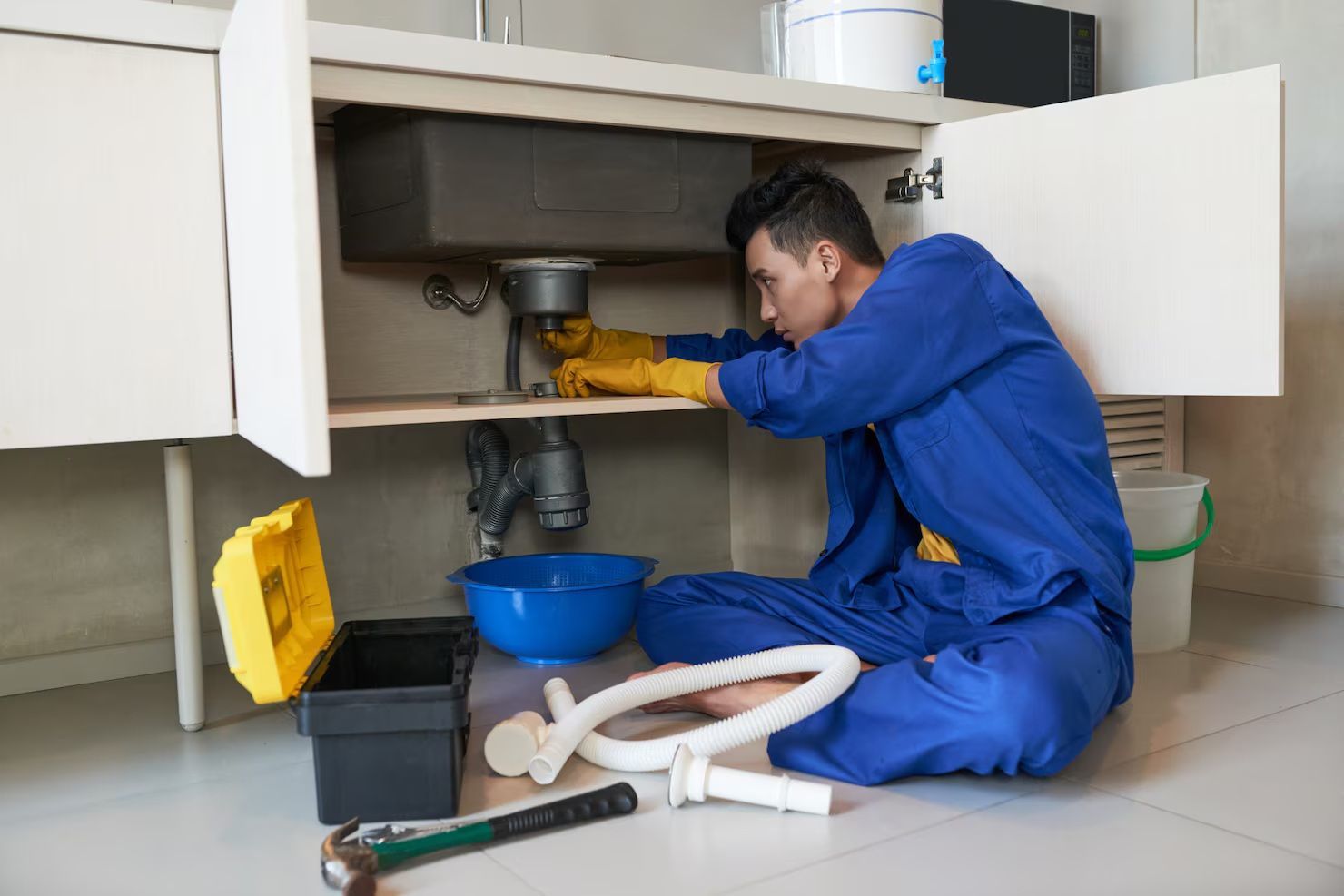 A technician in a blue uniform repairing sink plumbing under a cabinet with a toolbox and tools on the floor.