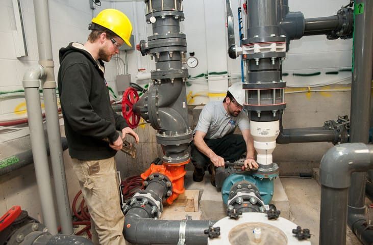 Two men are working on a machine in a room with pipes.