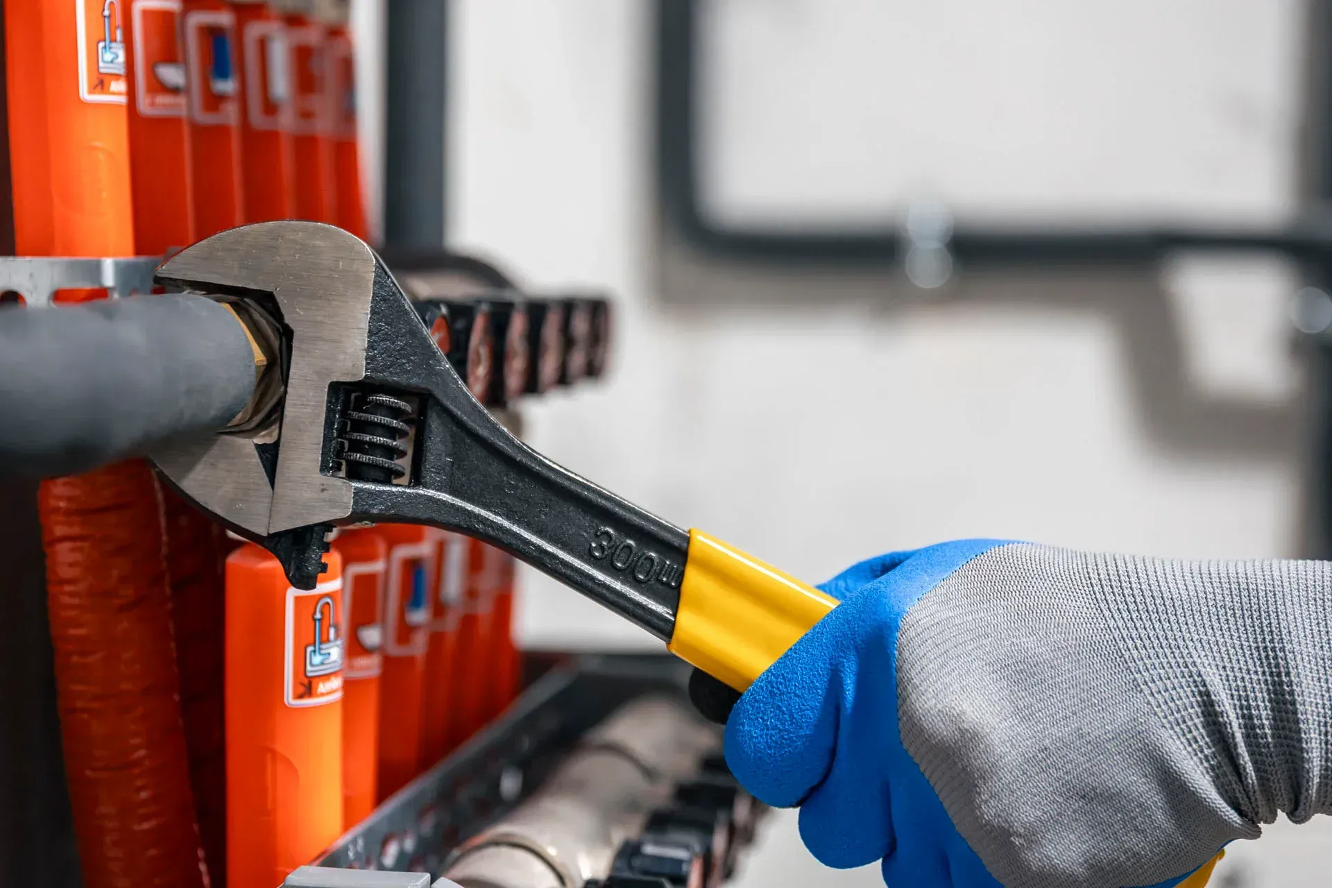 A gloved hand uses an adjustable wrench to tighten a fitting on an insulated pipe in a mechanical setting.