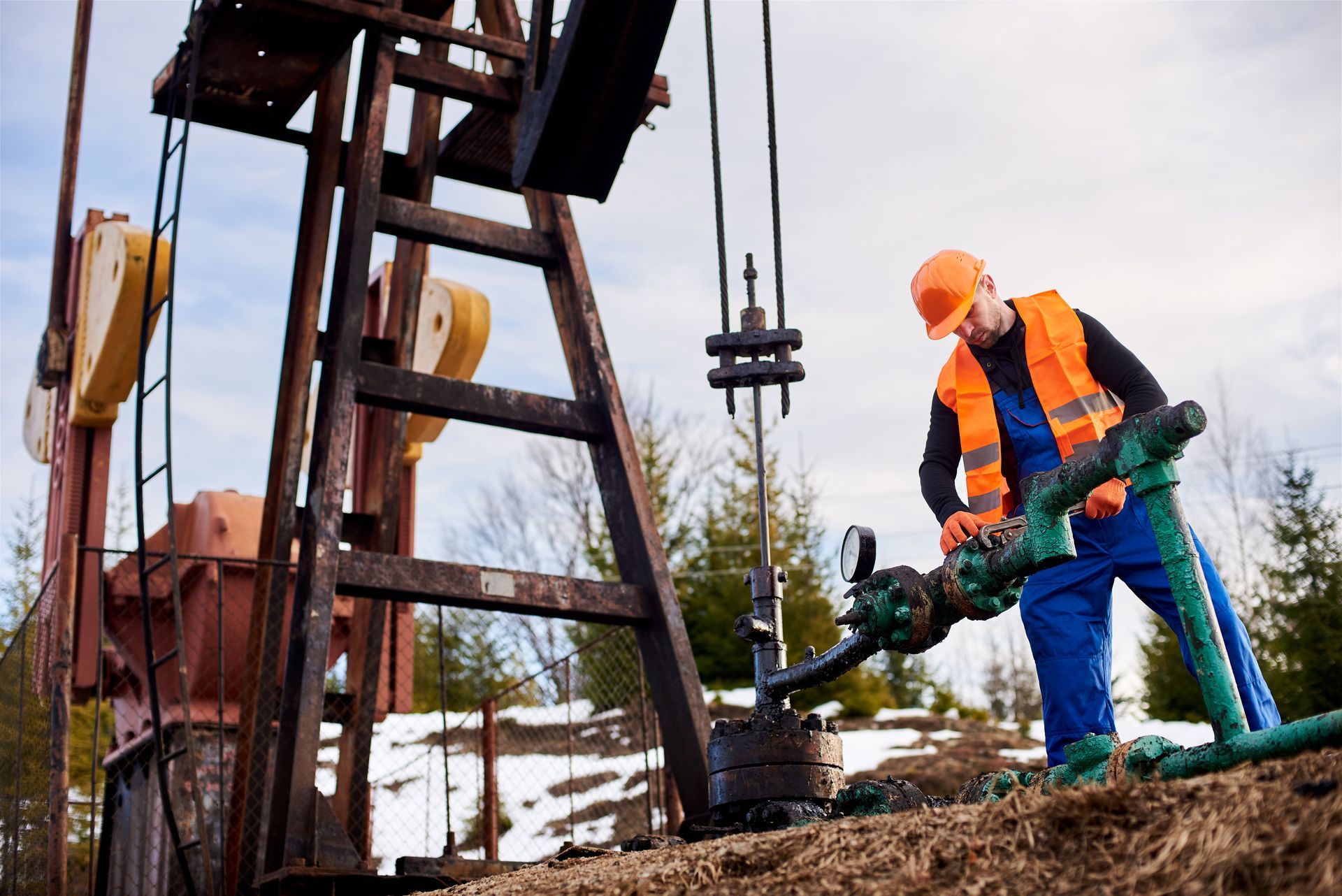 An oil industry worker in an orange vest and hard hat inspects pipes at an oil pumpjack in a rural, snowy setting.