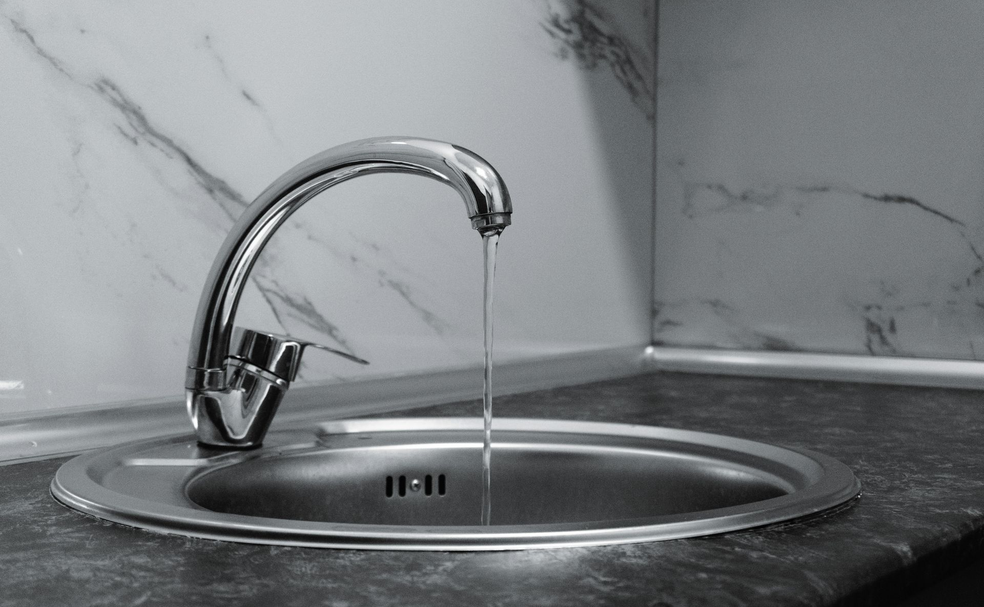 A silver kitchen faucet dripping a thin stream of water into a stainless steel sink against a marble-patterned backsplash.