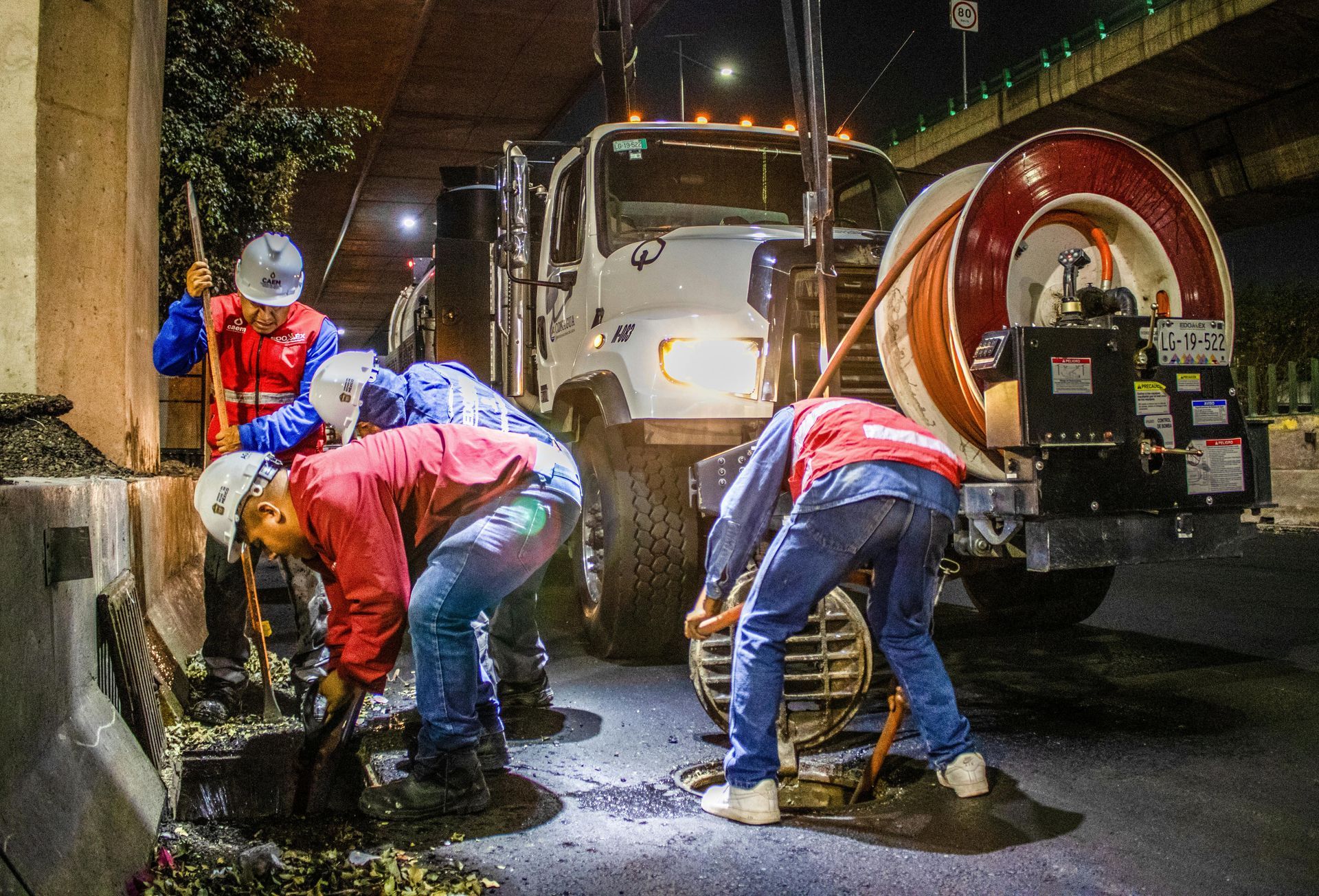 Workers in hard hats and red uniforms clean an urban storm drain at night near a utility truck with a large hose reel.