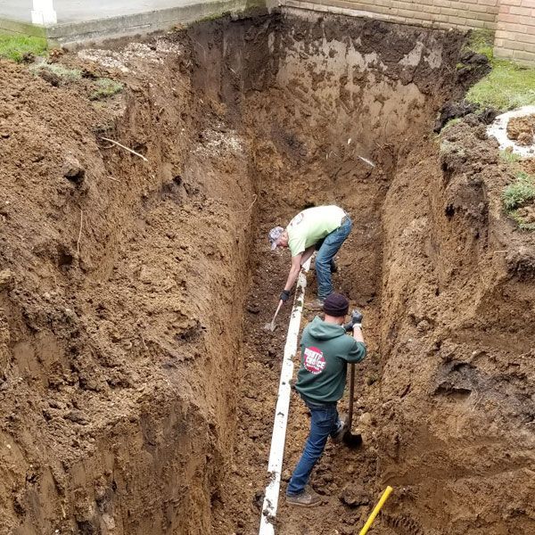 Two men are digging a hole in the ground to install a drain pipe.