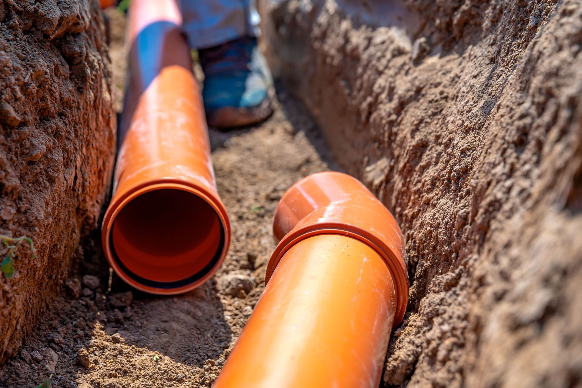 Orange PVC drainage pipes lie inside a narrow, freshly dug dirt trench.