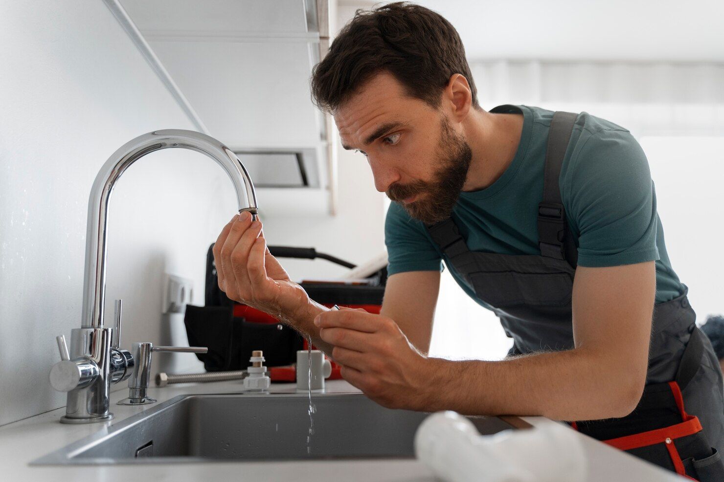 A plumber in a gray uniform inspecting a kitchen faucet over a sink while water drips from it.