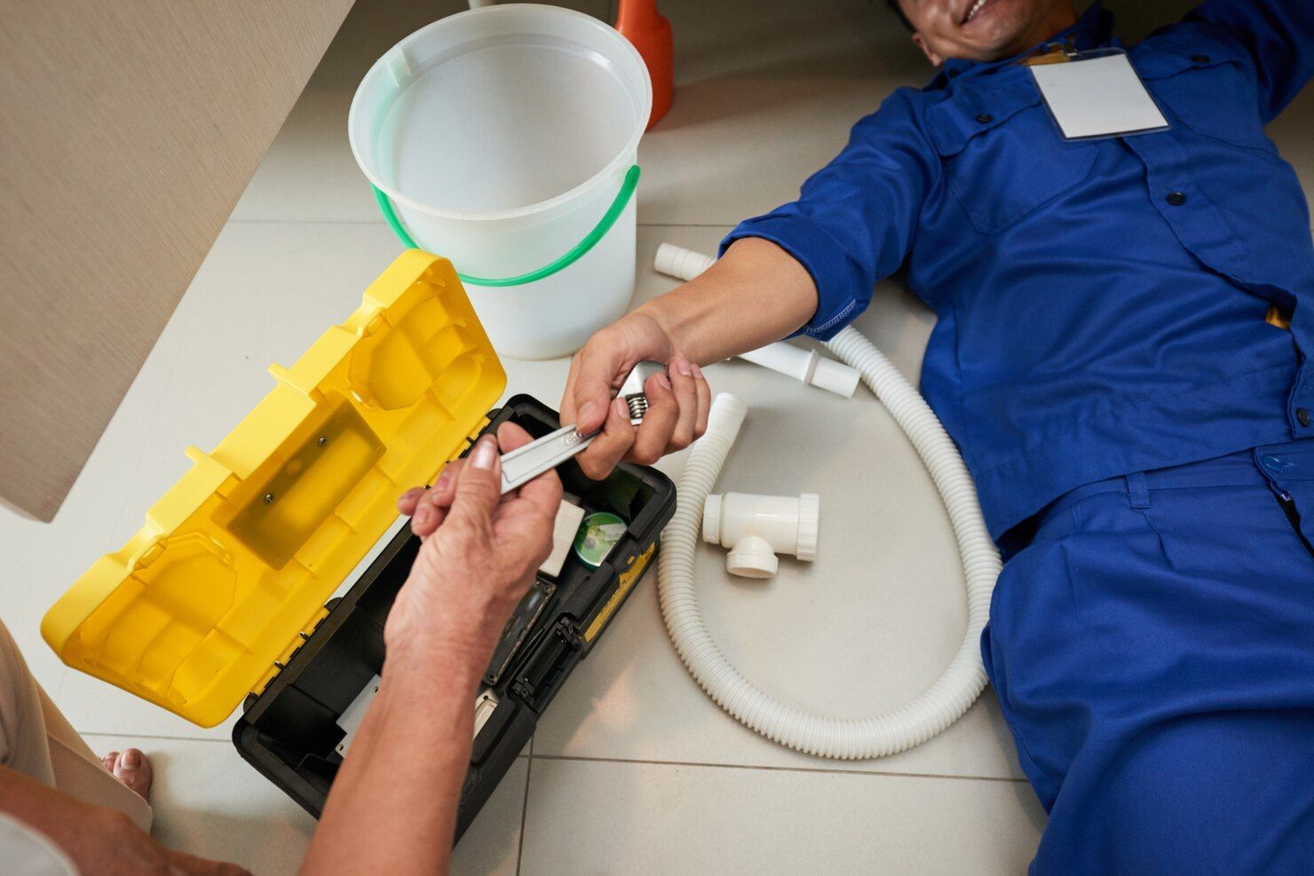 A plumber in a blue uniform lies on the floor, reaching out to accept a tool from someone holding an open yellow toolbox.