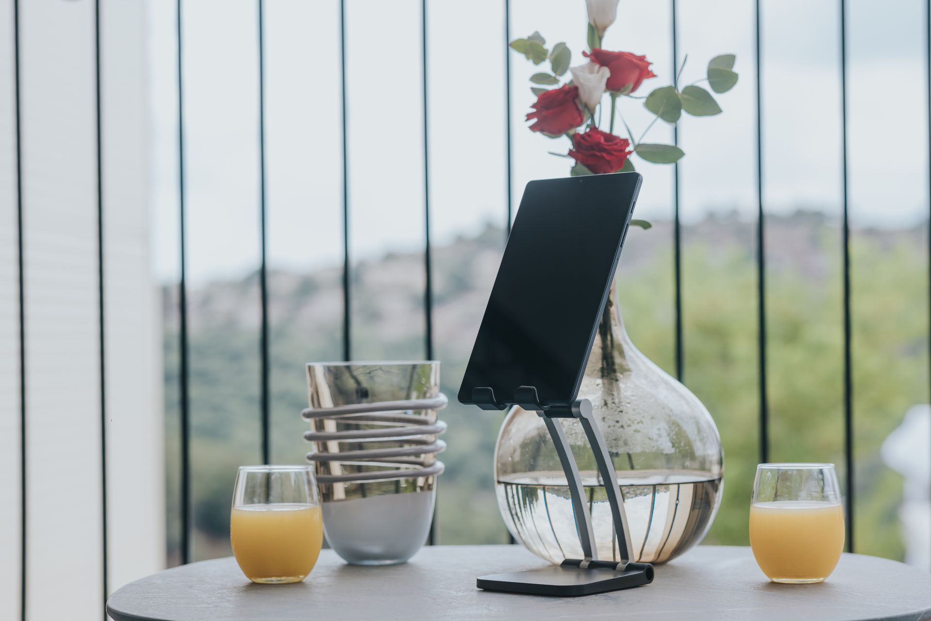 A tablet on a stand next to a vase with roses and two glasses of orange juice on a round table.