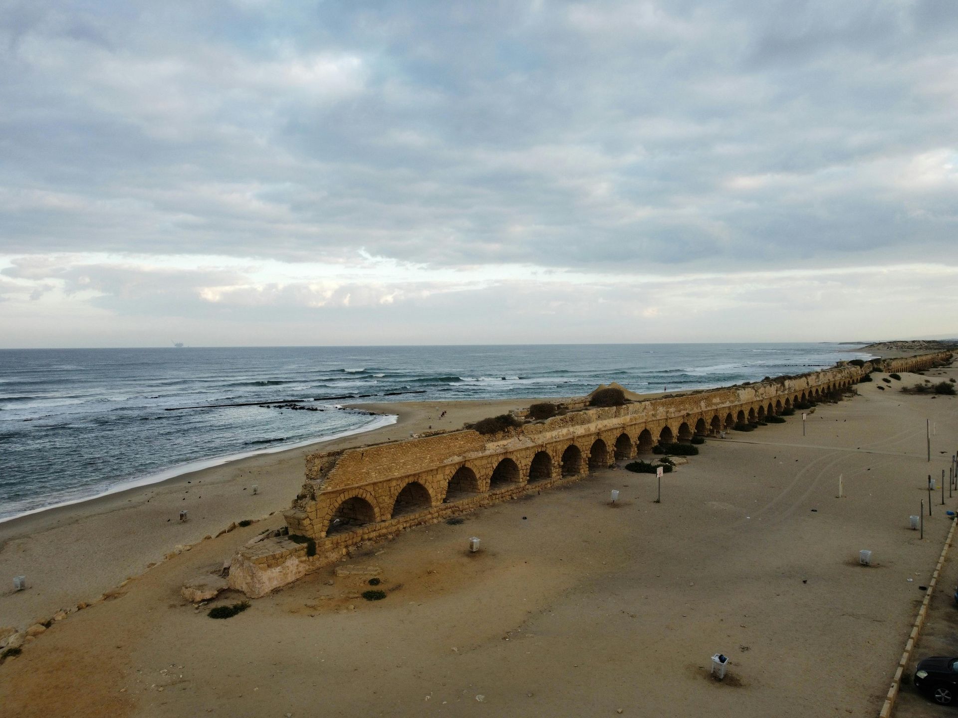 Ruined Roman aqueduct on a sandy beach, with ocean waves under a cloudy sky.