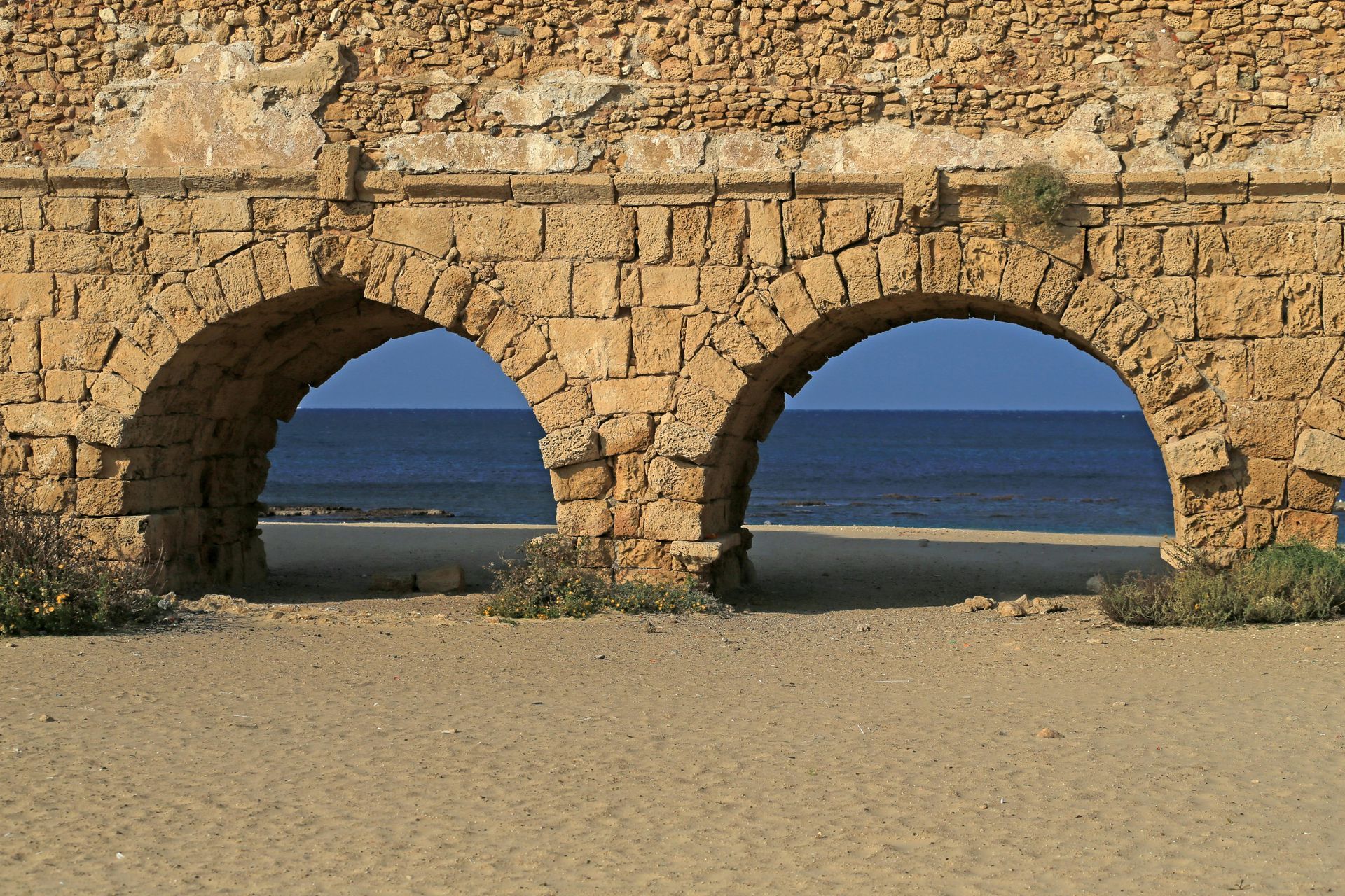 Arched stone aqueduct over a sandy beach, framing the blue sea and sky.