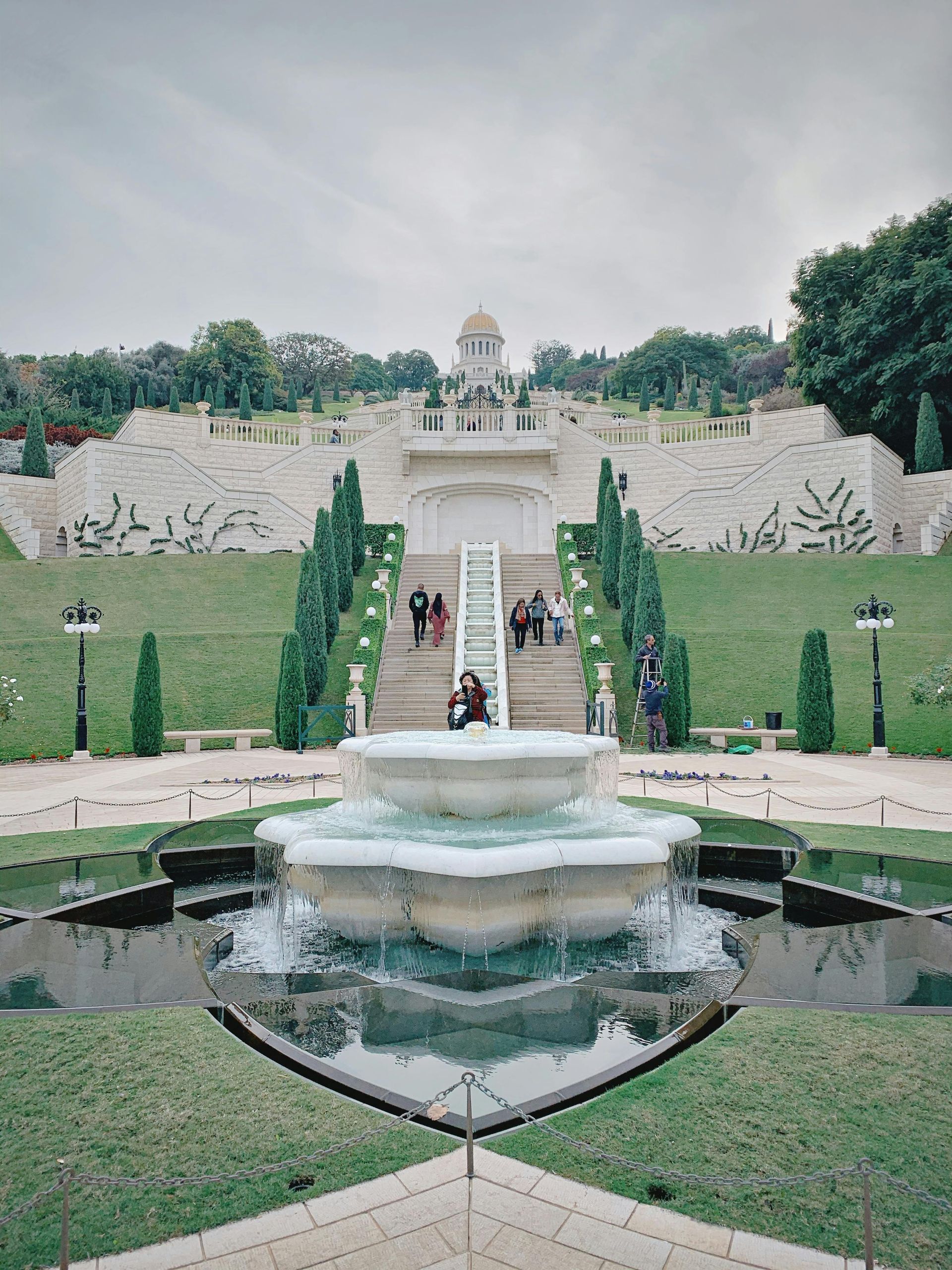 A tiered garden with a large fountain in the foreground, leading up to a white structure, stairs, and greenery.