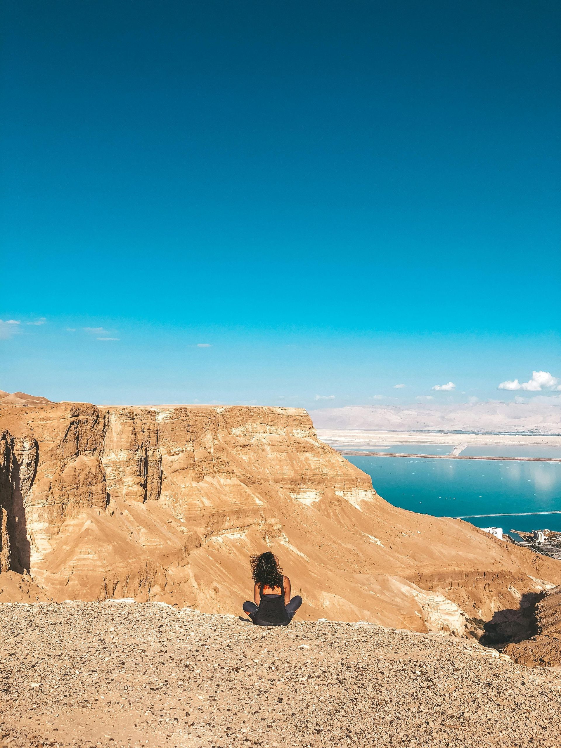 Woman sits on cliff overlooking water and landscape under blue sky.