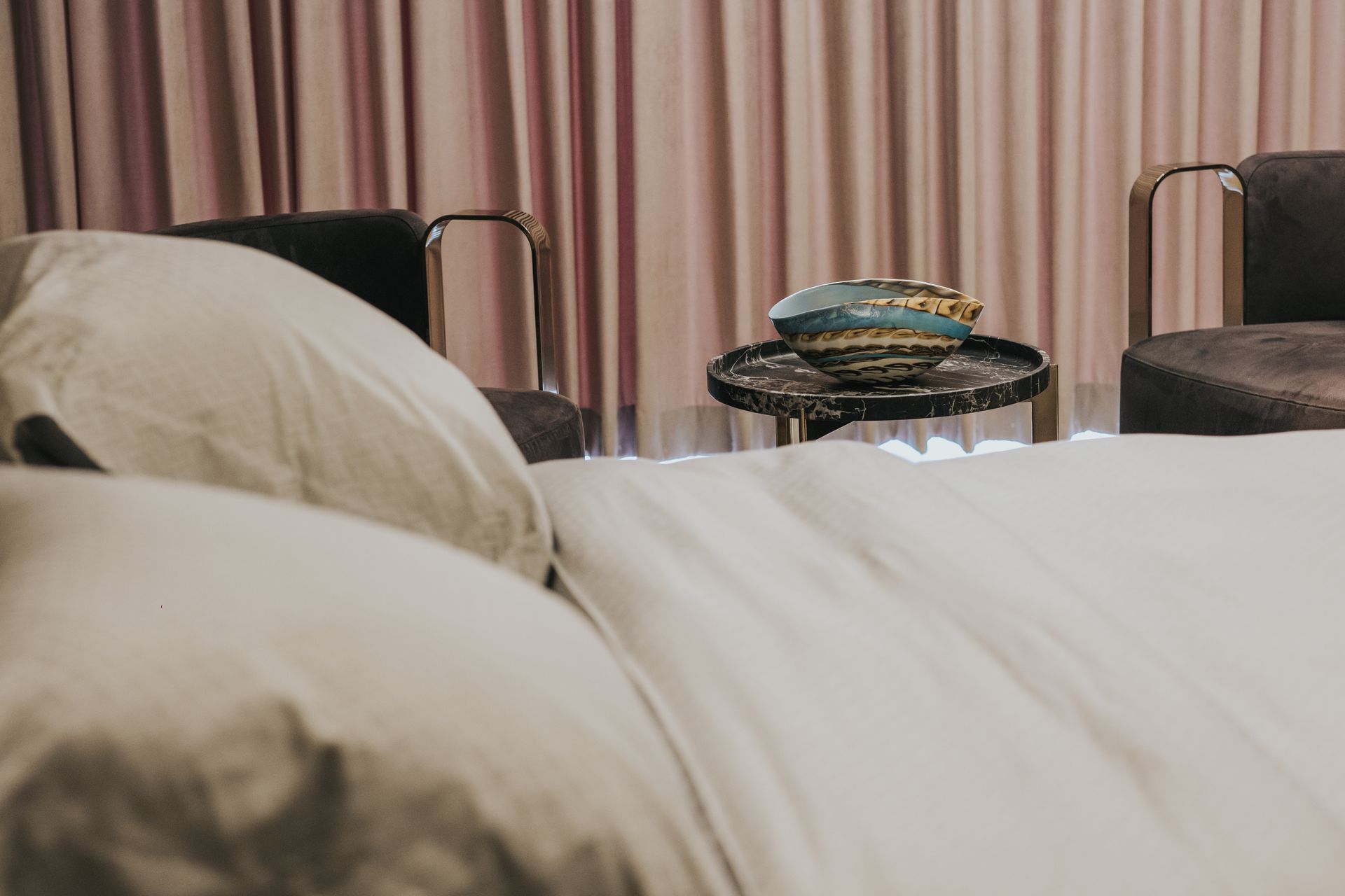Bedroom with white linens, two chairs, and a small table with an ornament against pink curtains.