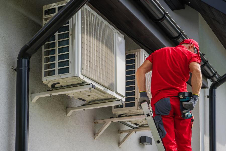 A man is standing on a ladder fixing air conditioners on the side of a building.