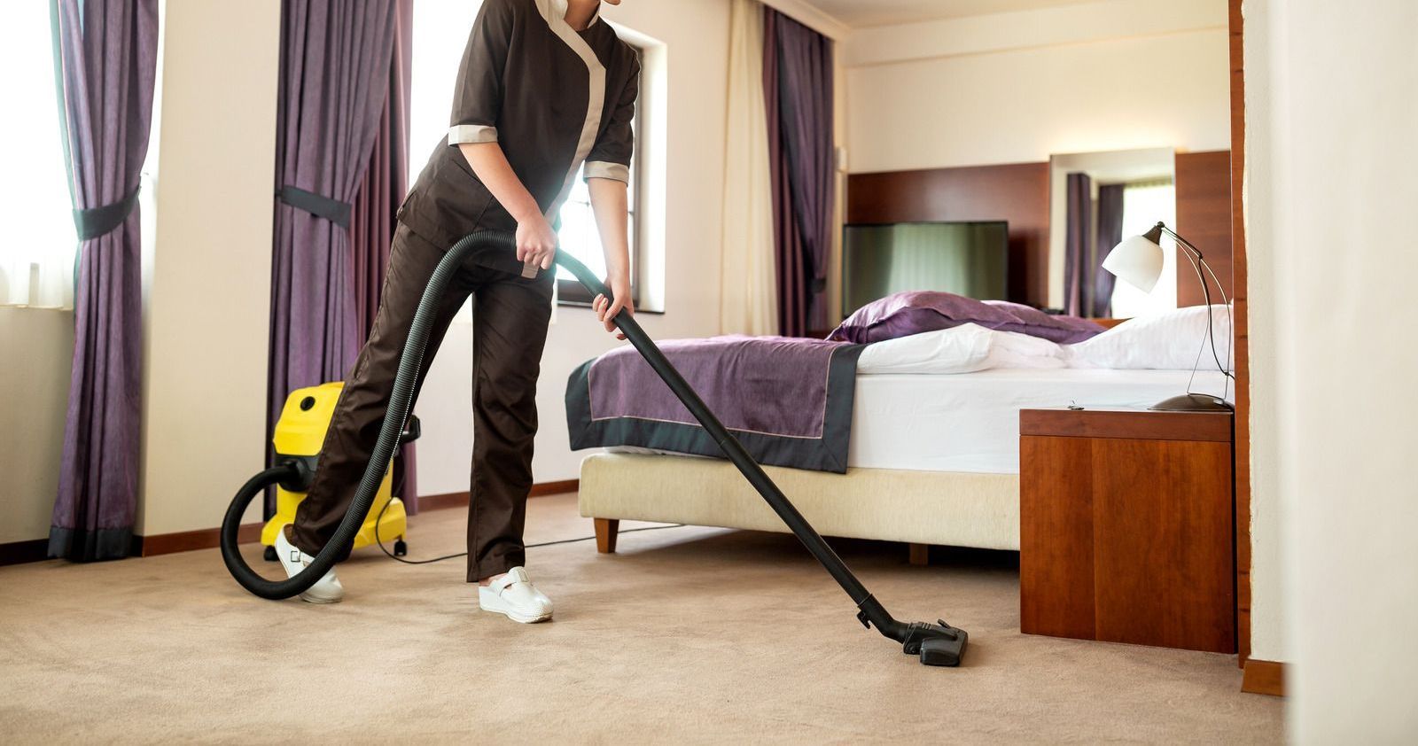 Hotel housekeeper vacuuming a carpeted bedroom with a yellow vacuum cleaner.