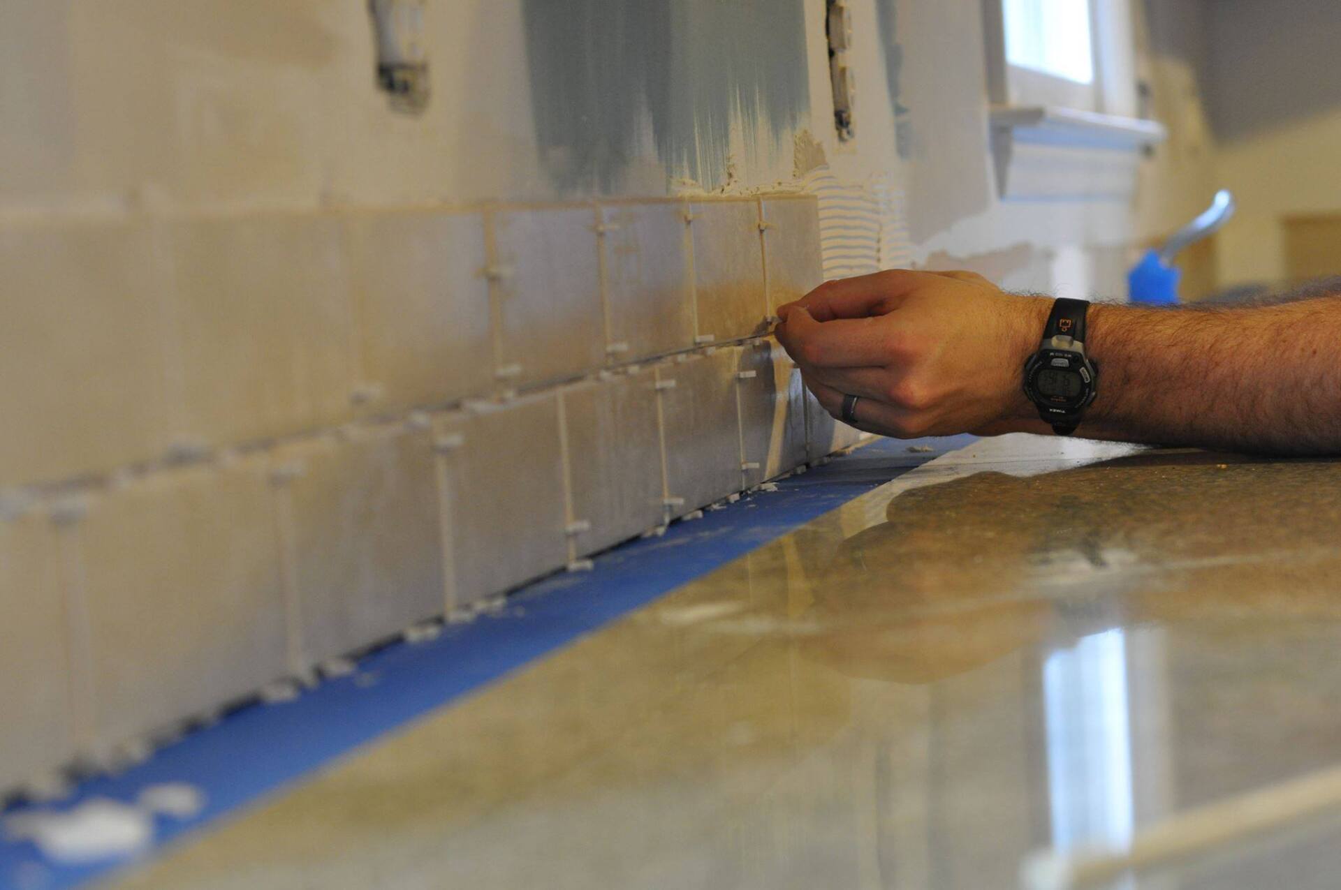 A man is applying glue to a tile wall in a kitchen.