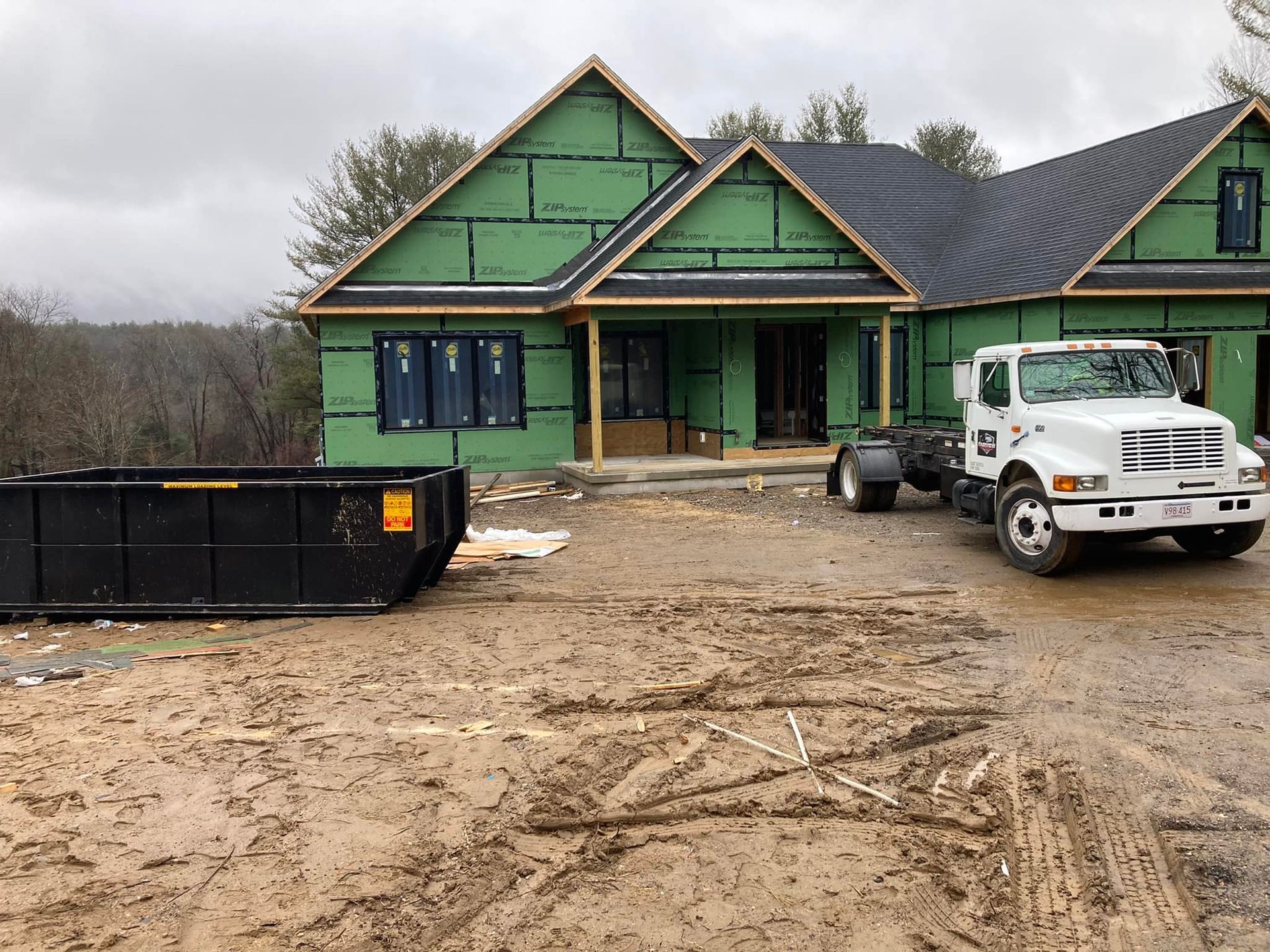 New house under construction, muddy ground, green siding, black dumpster, white truck parked.