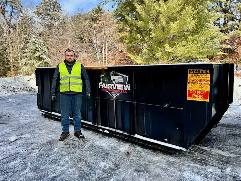 A man stands next to a large black dumpster in a snowy outdoor setting.