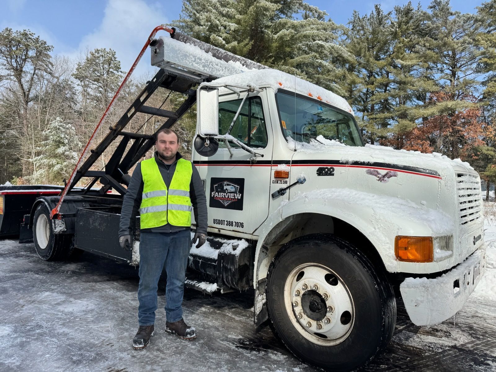 Man in reflective vest stands beside a white snow-covered truck with a dumpster lift in a snowy area.