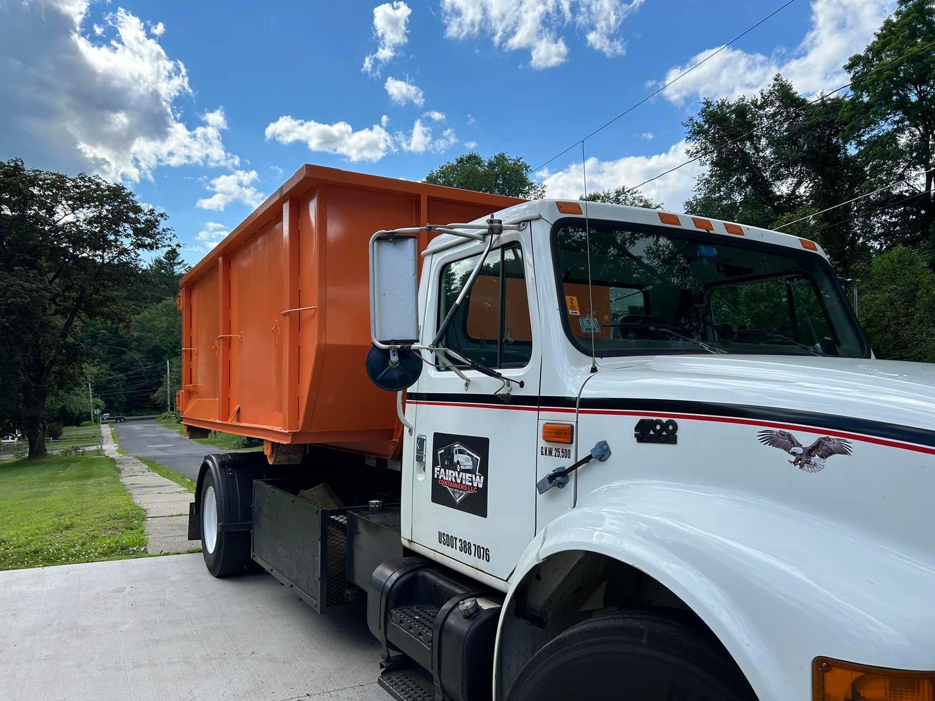Orange dumpster on a white truck on a residential street under a blue sky with clouds.