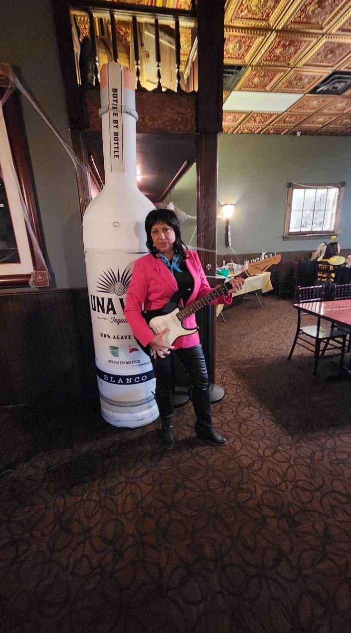 A woman is standing in front of a giant bottle of vodka holding a guitar.