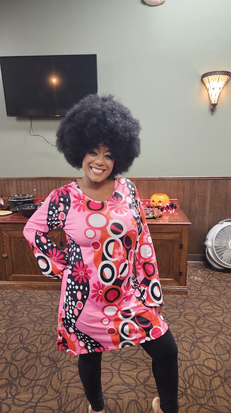 A woman wearing an afro wig and a pink dress is standing in a room.