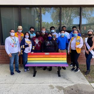 A group of people wearing face masks are standing around a rainbow bench.