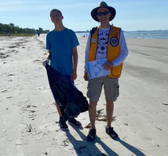 Two men standing on a beach one wearing a vest that says lions on it