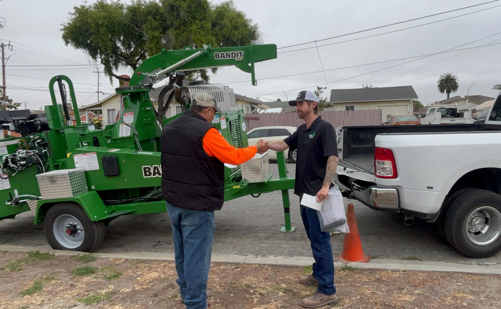 Two men shaking hands beside a green wood chipper and a white truck. Outdoor setting.