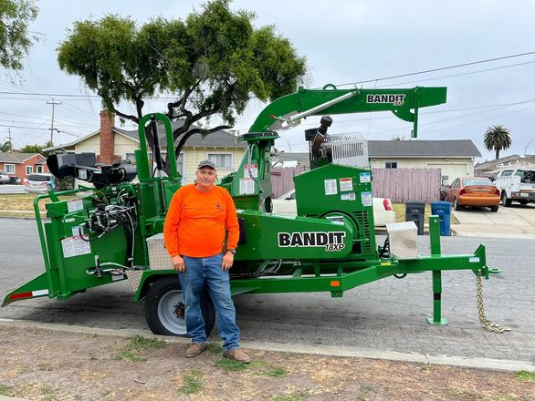 Man in orange shirt stands beside a green Bandit wood chipper on a street.