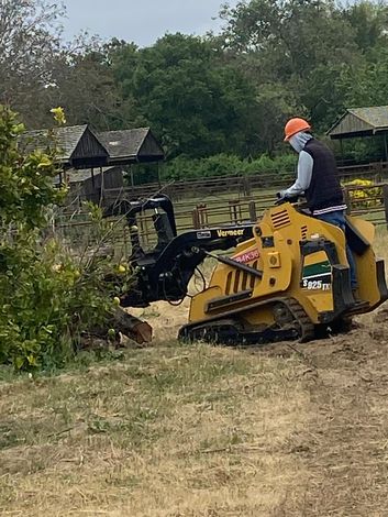 Man operating a yellow skid steer to clear brush outdoors, wearing a helmet.