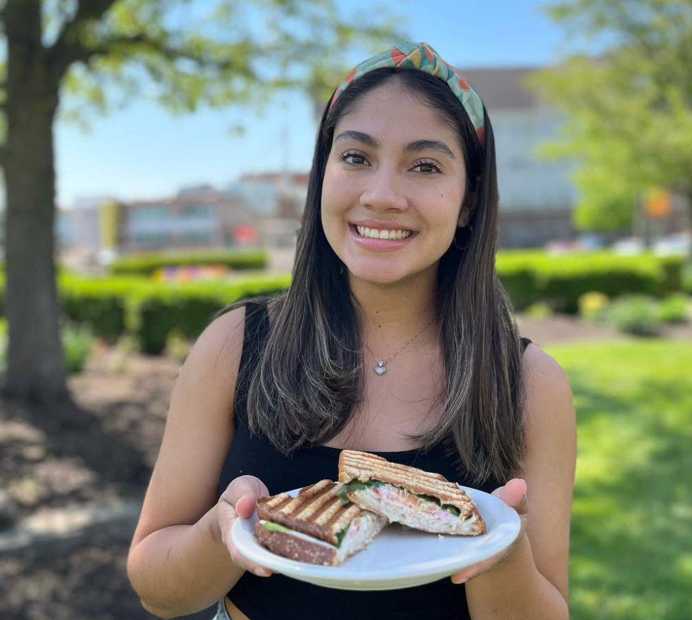 Woman holding a plate with a grilled sandwich, smiling outside on a sunny day.